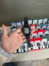 Hand holding a black luxury men's watch with a skeleton dial, multiple boxed watches displayed in the background, Ministry of Mens Affair premium collection.
